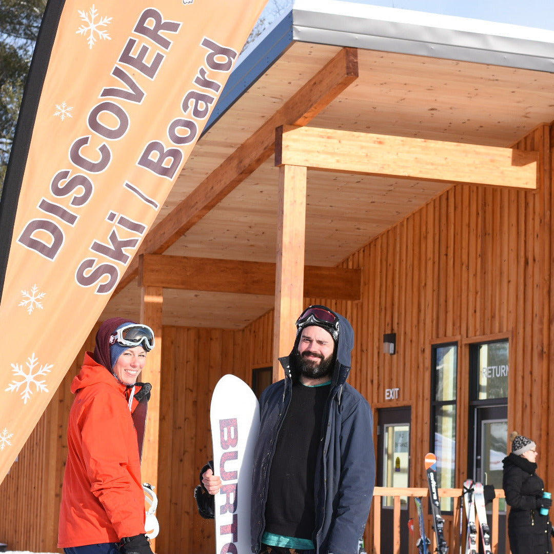 People with ski equipment in front of a wooden building with a 'Discover Ski Board' sign.