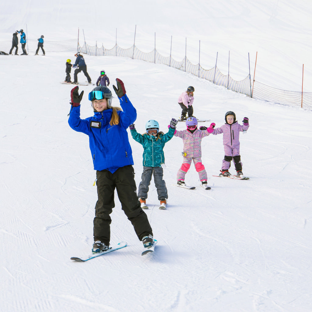 A group of children and an adult on a snowy mountain slope, with the adult in a bright blue ski jacket and black pants standing on a snowboard in the foreground while others ski behind them.