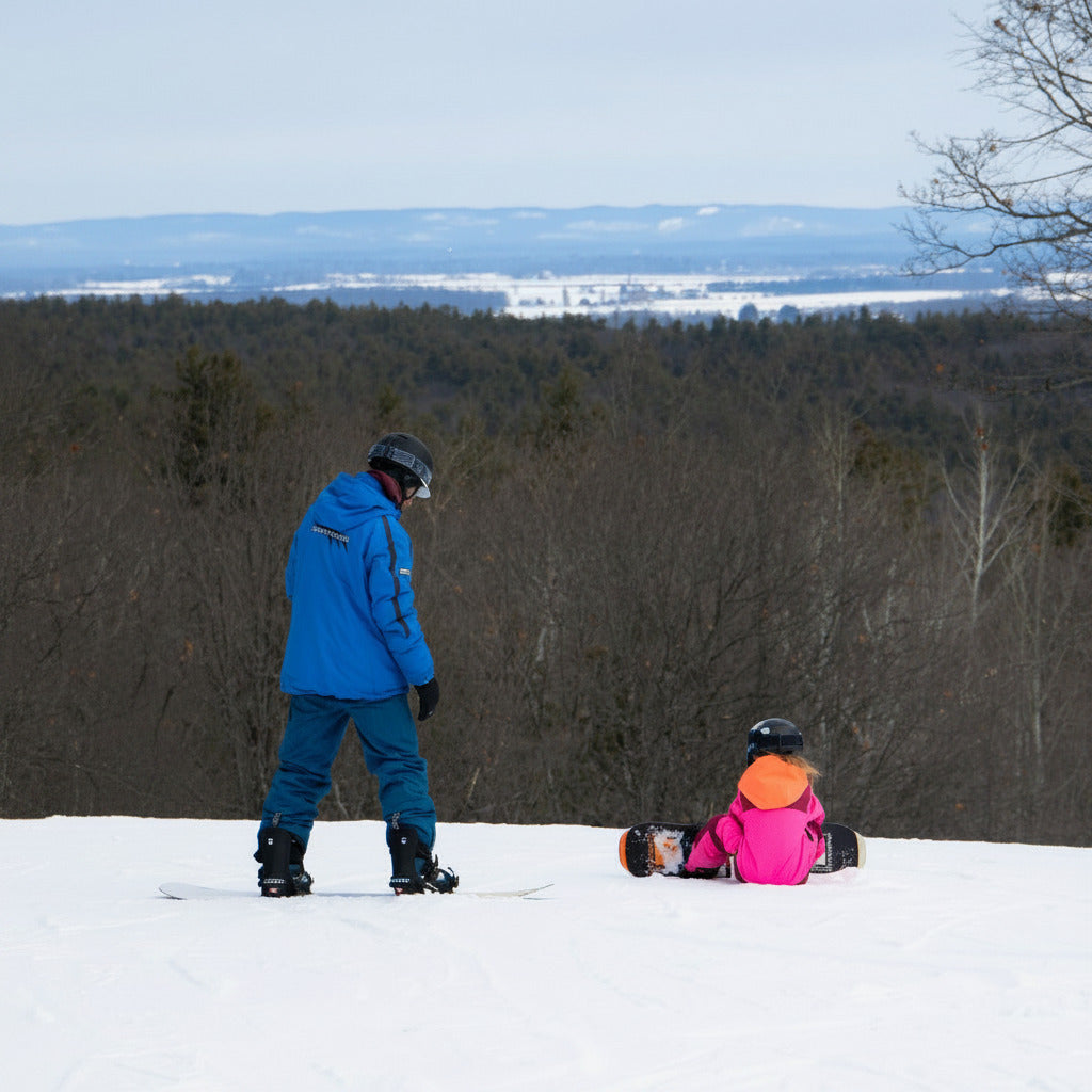 A snowy outdoor ski resort scene with three people on snowboards or sitting on the snow, with bare trees and distant hills under a pale sky.