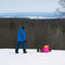 A snowy outdoor ski resort scene with three people on snowboards or sitting on the snow, with bare trees and distant hills under a pale sky.