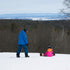 A snowy outdoor ski resort scene with three people on snowboards or sitting on the snow, with bare trees and distant hills under a pale sky.