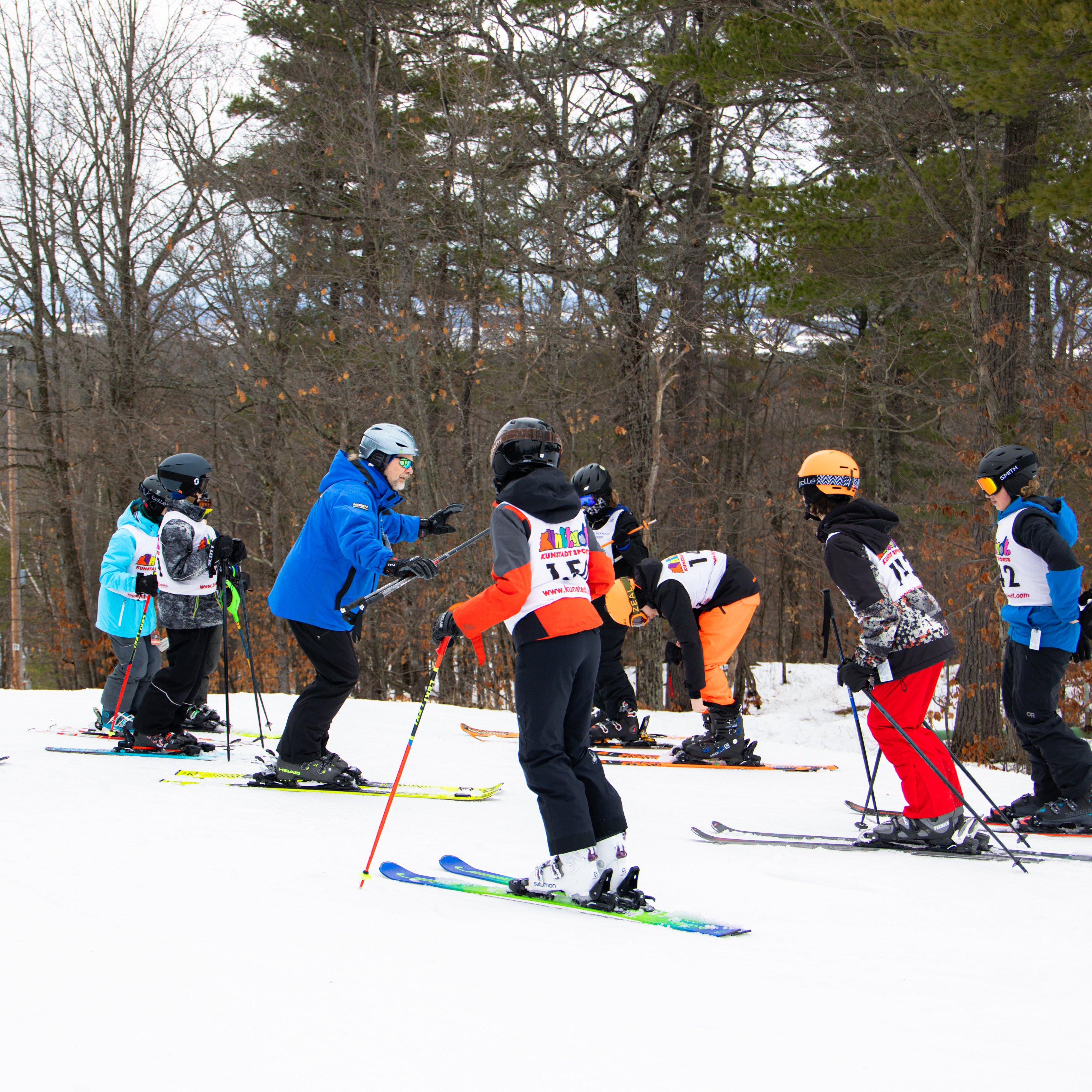 A group of children wearing colorful winter sports gear, helmets, and skis standing on a snowy slope with bare trees in the background.