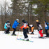 A group of children wearing colorful winter sports gear, helmets, and skis standing on a snowy slope with bare trees in the background.