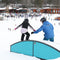 A person in a black winter outfit balances on a turquoise snowboard ramp while being assisted by another person in a blue jacket on a snowy mountain slope with ski resort buildings and other skiers in the background.