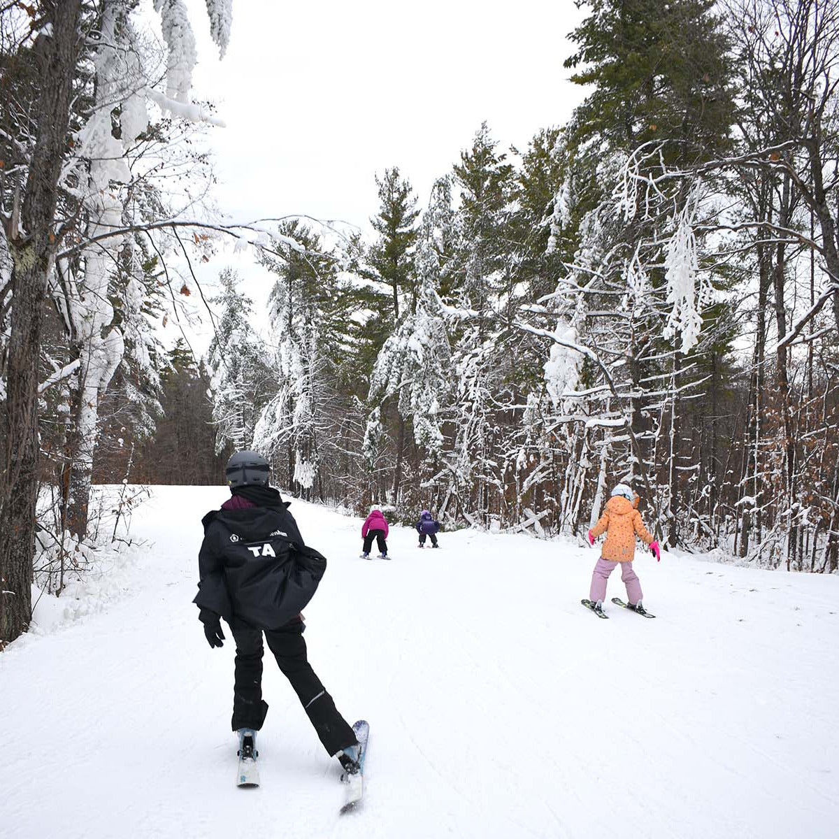Youth ski and snowboard program with adults assisting children on a ski slope with snow-covered trees in the background.