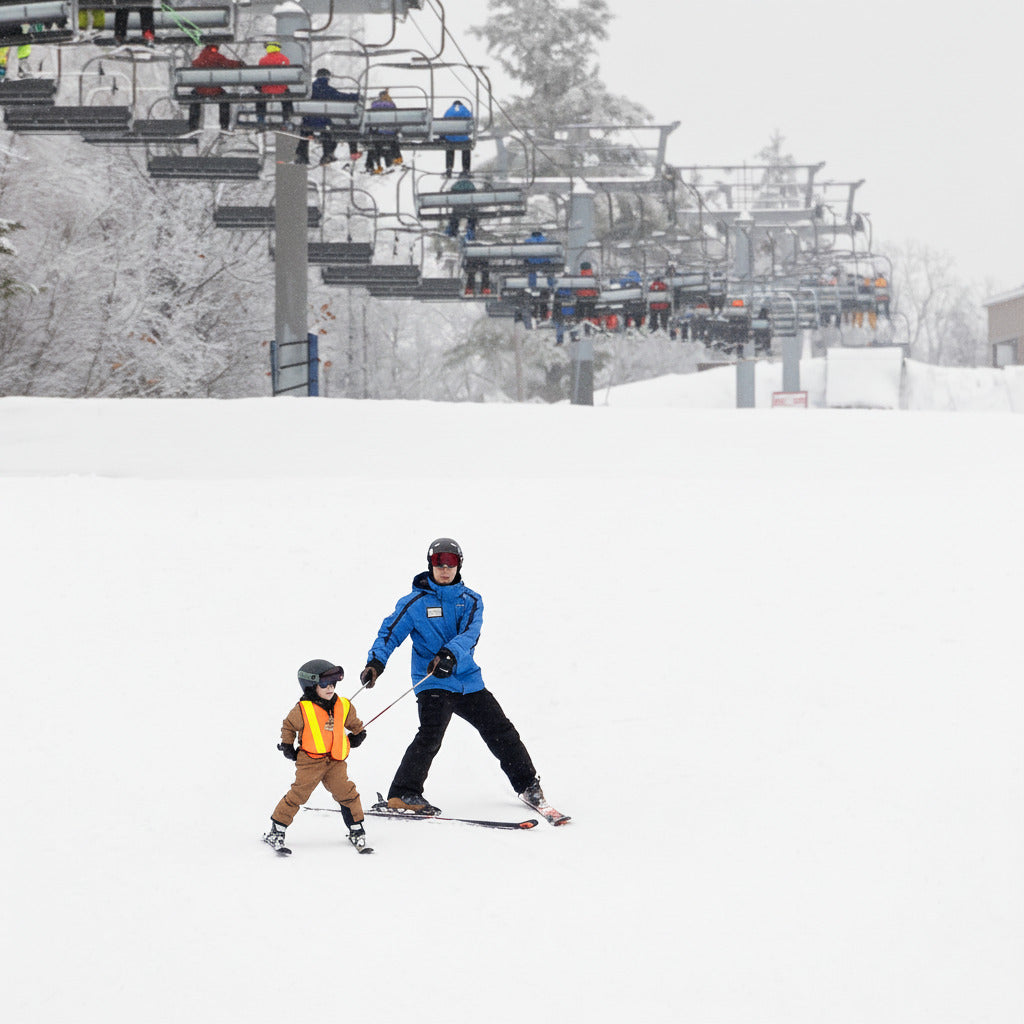 An adult and a child skiing down a snow-covered slope with ski lifts in the background.