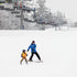 An adult and a child skiing down a snow-covered slope with ski lifts in the background.