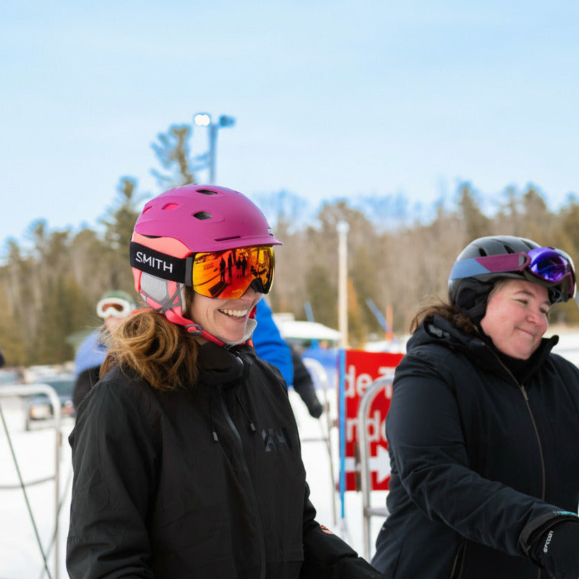 Two smiling women on a ski resort wearing helmets, goggles, and ski jackets, with trees and other skiers in the background.