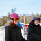 Two smiling women on a ski resort wearing helmets, goggles, and ski jackets, with trees and other skiers in the background.