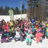 A group of children and adults at a ski resort, dressed in ski gear, posing for a group photo on the snow-covered ground.