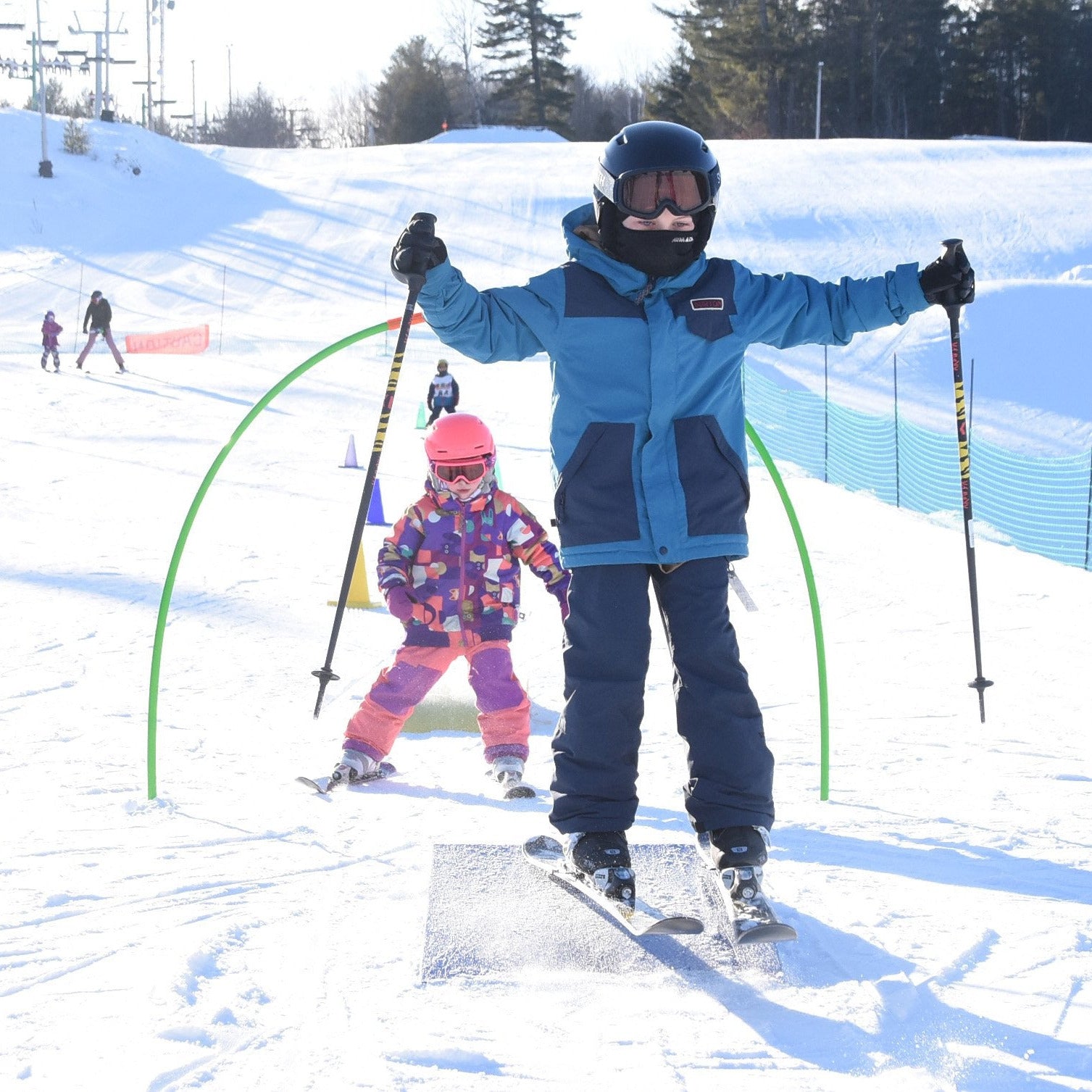 Two children on a ski resort slope, dressed in ski gear, posing with their skis and poles. The child in the foreground is smiling and extending their arms, while the other child is slightly behind and to the side, also with a happy expression.