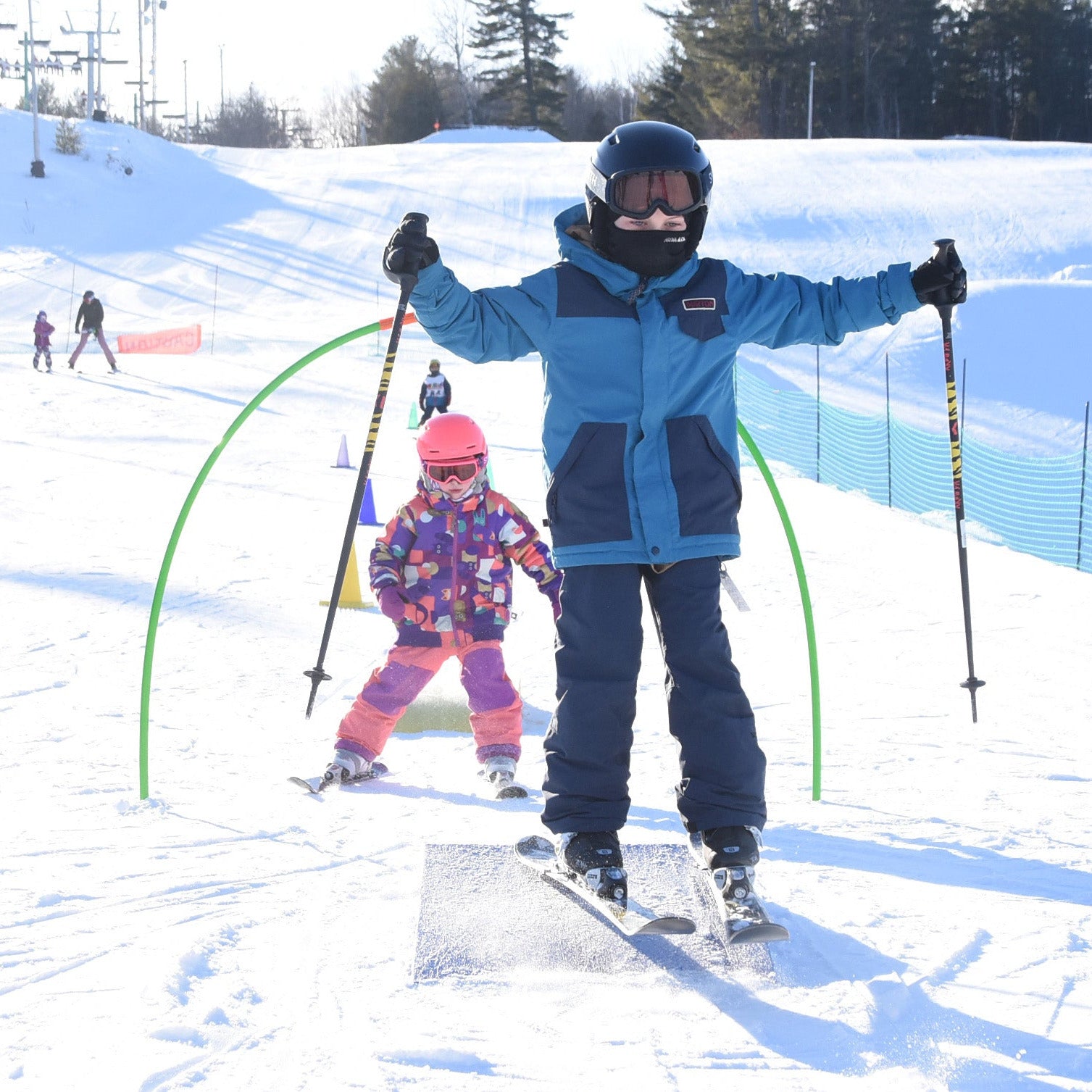 Two young children, both wearing ski gear, standing on skis and snowboards respectively, with arms raised. They are on a snow-covered slope with other skiers in the background.