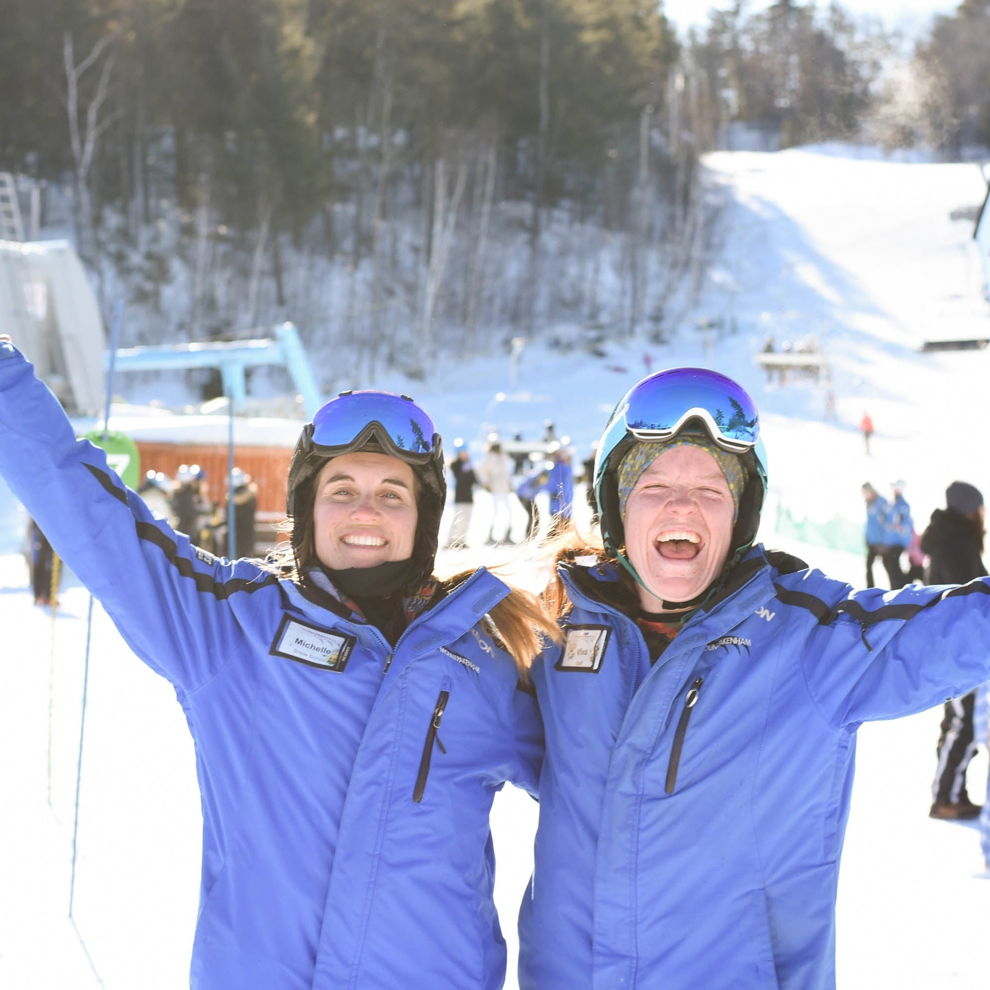 Two adults in ski gear, standing with arms raised and smiling, on a snow-covered mountain with ski lifts and other skiers in the background.