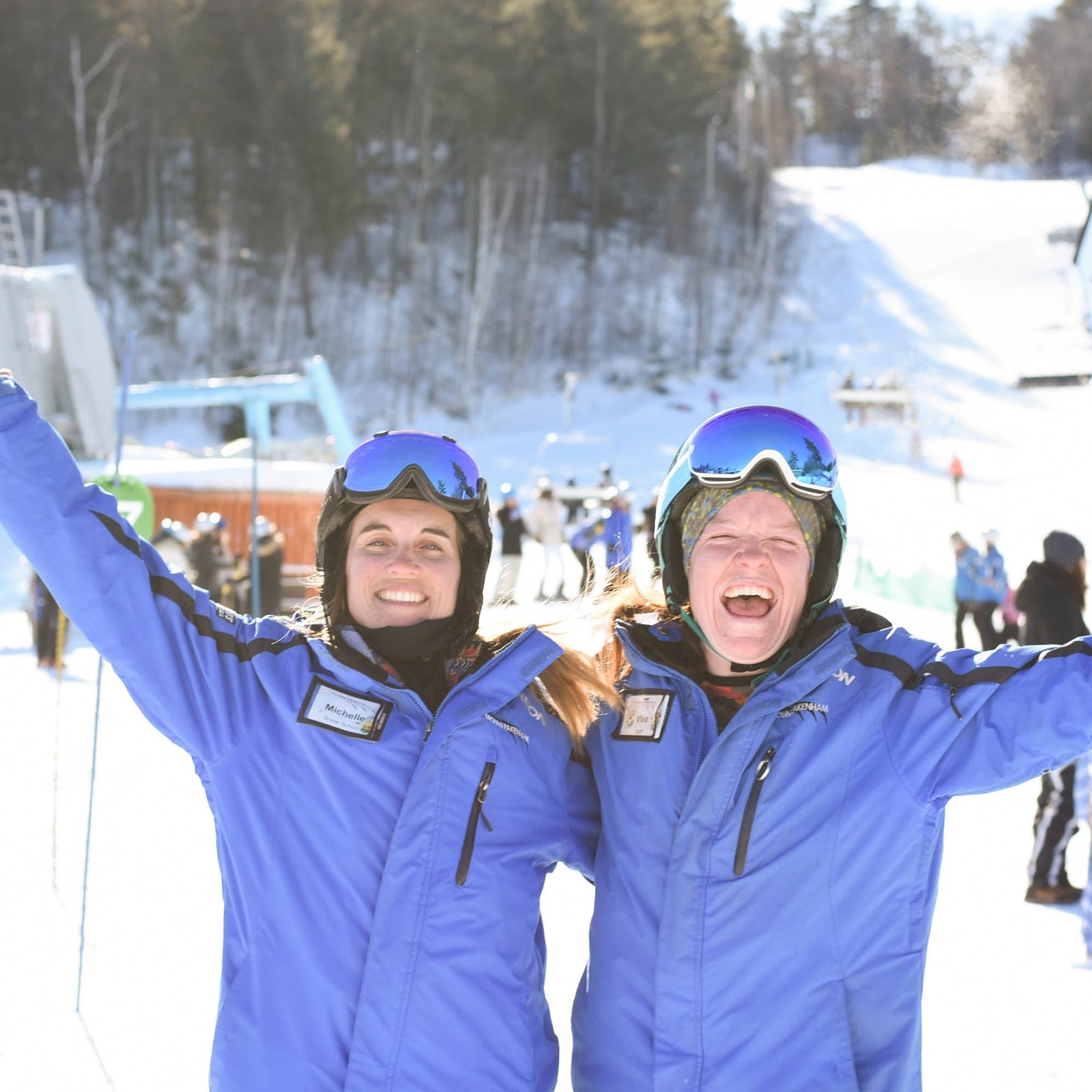 Two adults in ski gear, standing with arms raised and smiling, on a snow-covered mountain with ski lifts and other skiers in the background.