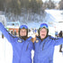 Two adults in ski gear, standing with arms raised and smiling, on a snow-covered mountain with ski lifts and other skiers in the background.